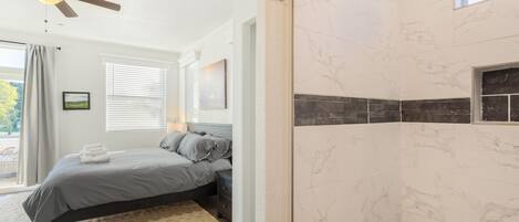 A bedroom with a bed, a ceiling fan, a window with blinds, and a bathroom with marble tiles.