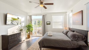 Quarto com cama, ventilador de teto, cômoda, planta e janela com persianas.