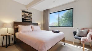 Ein Schlafzimmer mit einem großen Bett, einem Nachttisch mit Lampe, einem Sessel mit Decke und einem Fenster mit Blick auf Bäume.