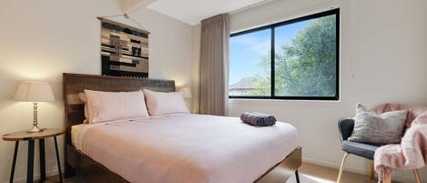 A bedroom with a large bed, a bedside table with a lamp, a chair with a blanket, and a window with a view of trees.