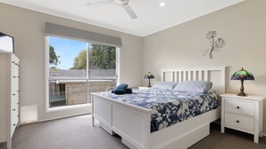 A bedroom with a bed, two bedside tables, a ceiling fan, a window with blinds, and a decorative wall piece. - Blue Bayou in Coronet Bay (Coronet Bay)