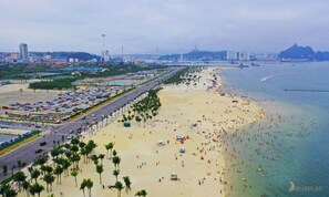 A beach with a long promenade lined with palm trees, a parking lot, and a view of the sea and distant mountains. - Villa Royal Lotus Ha Long ( Standard villa, with shared swimming pool) (Hạ Long)