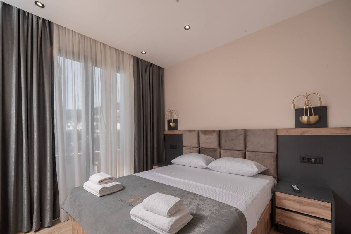 Ein modernes Hotelzimmer mit Bett, Nachttischen, einem Schreibtisch und einem großen Fenster mit Vorhängen.