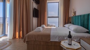 Habitación de hotel con una cama, una mesita de noche con una botella y un vaso, un televisor montado en la pared y un ventanal con cortinas.