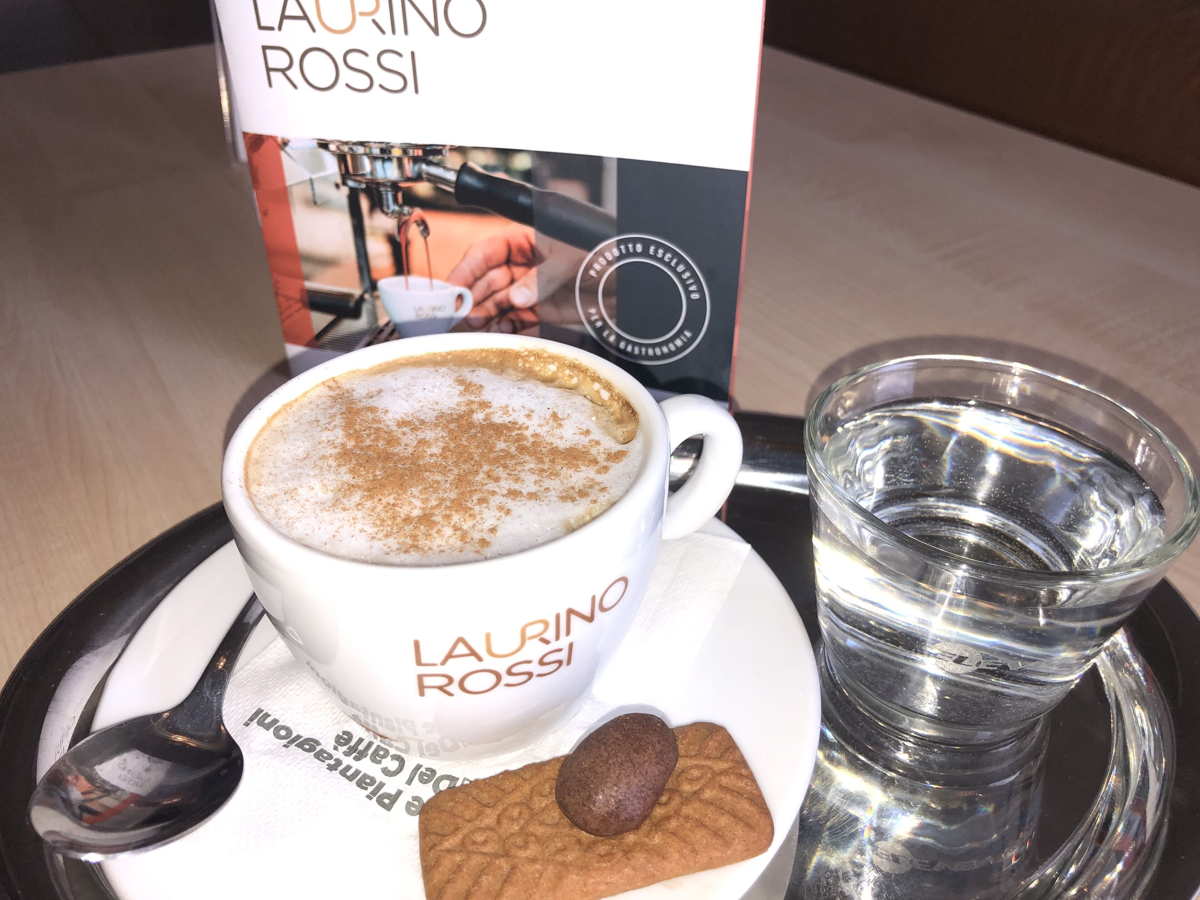 A cup of coffee with a biscotti on a tray, a glass of water, and a menu or brochure with the text "LAURINO ROSSI".