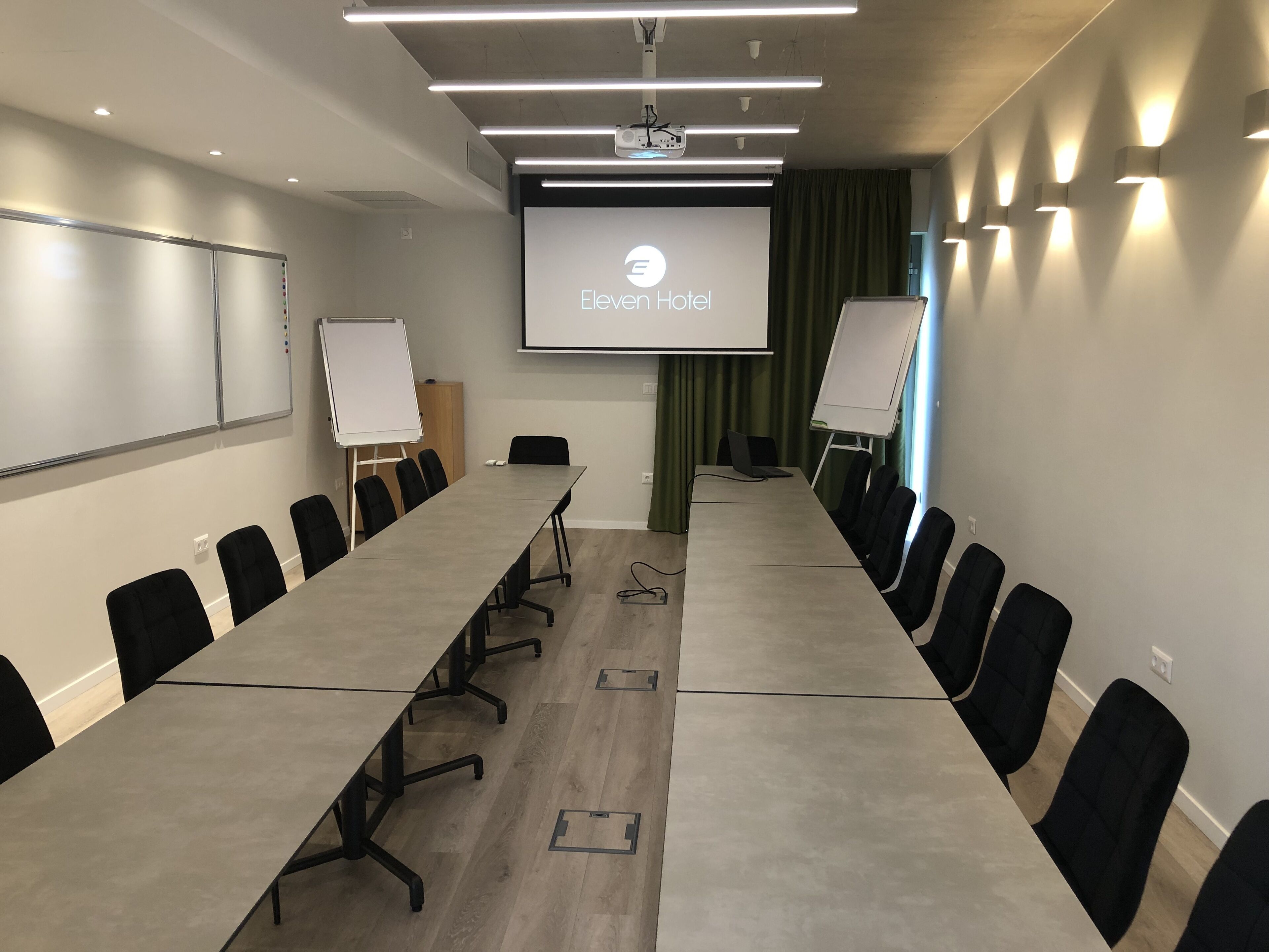 A conference room with a long table, black chairs, a whiteboard, a projector screen displaying the Eleven Hotel logo, and a whiteboard stand.
