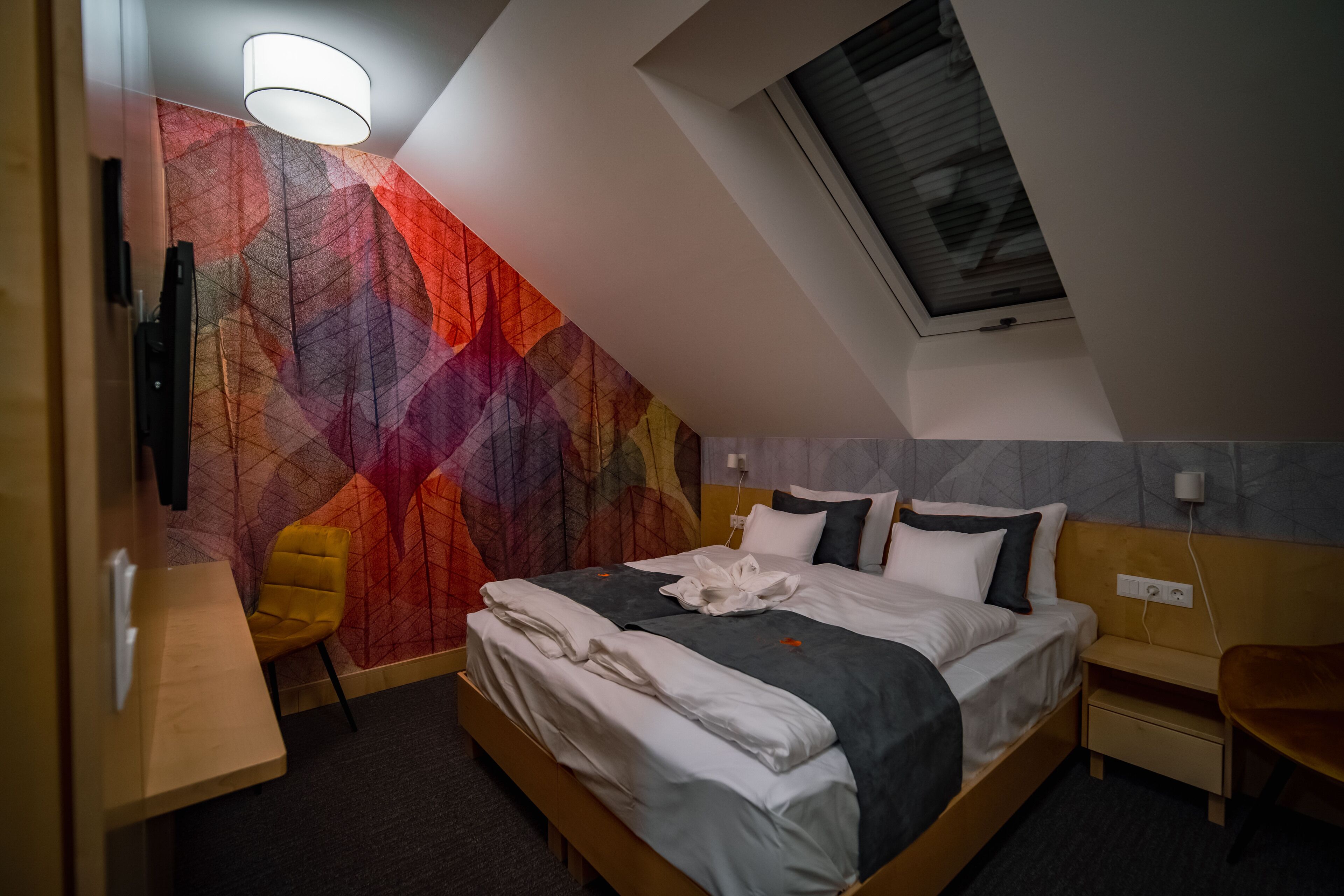 Una habitación de hotel moderna con techo inclinado, una cama grande con ropa de cama blanca y gris, una silla amarilla y un colorido mural abstracto.