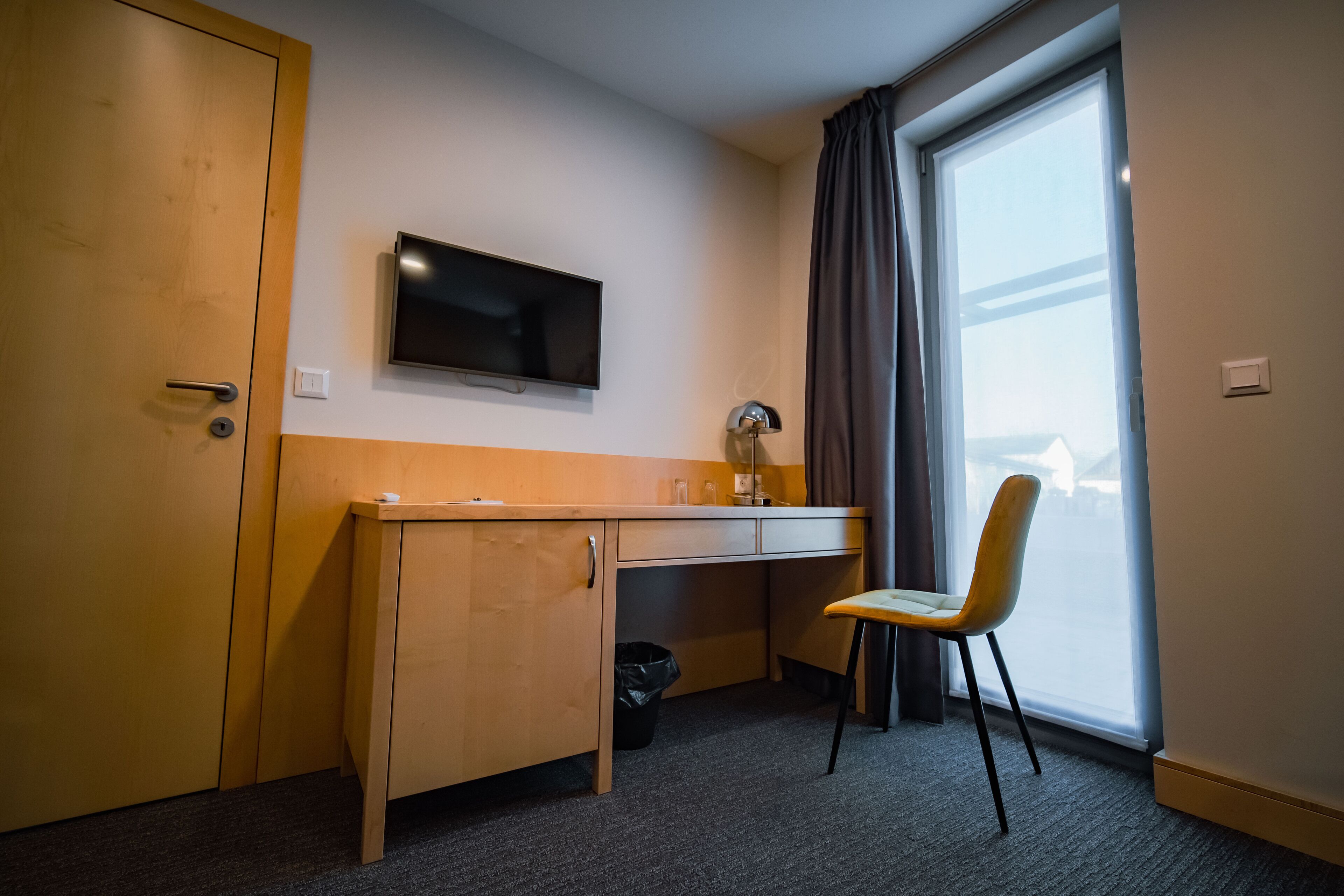Habitación de hotel con escritorio de madera, televisor montado en la pared, una silla y un ventanal con cortinas.