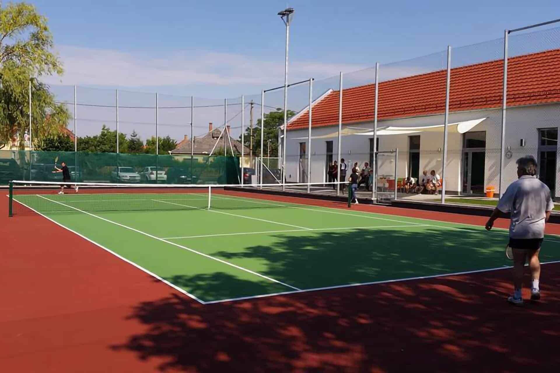 An outdoor tennis court with a net, surrounded by a fence and a building with a red roof.