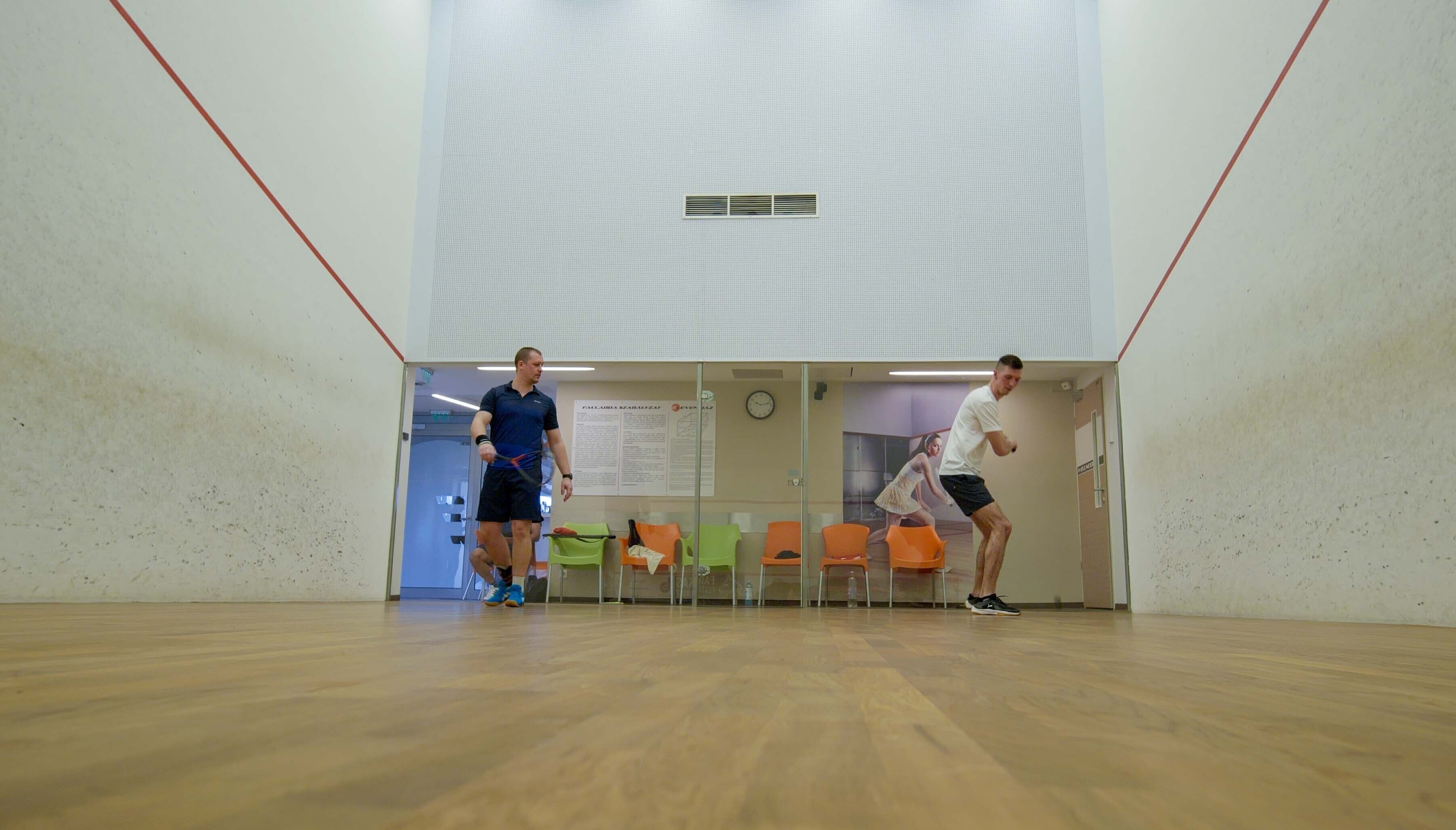 A squash court with wooden flooring, glass walls, and orange chairs.