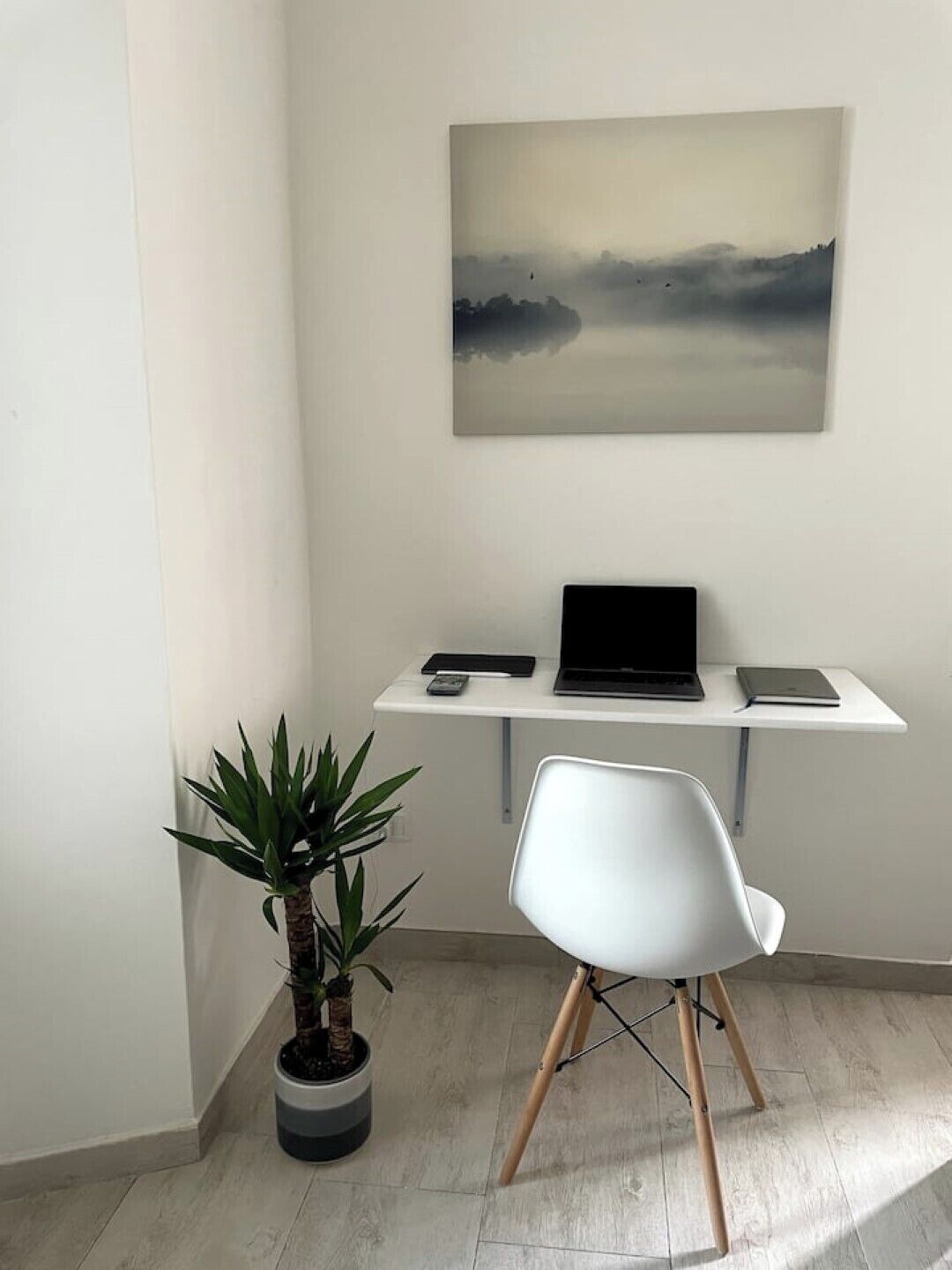 Um espaço de escritório minimalista com uma mesa branca, um laptop, um smartphone e um tablet. Há uma cadeira branca com pés de madeira e uma planta em vaso com folhas verdes.