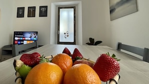 Ein Esstisch mit Obstkorb, ein Fernseher, der eine Streaming-Dienst-Oberfläche zeigt, eine Tür mit Milchglasfenster und ein Bild an der Wand.