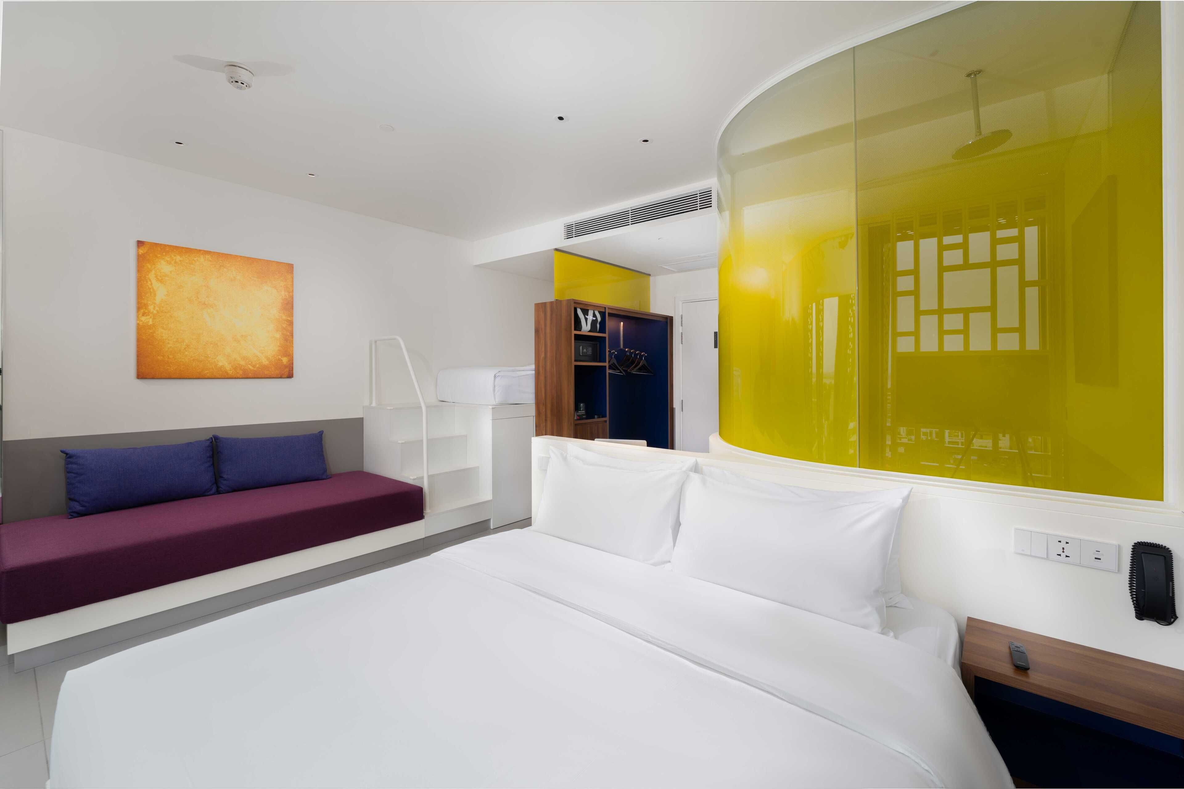 A modern hotel room with a large bed, a sofa, a wardrobe, and a bright yellow accent wall.