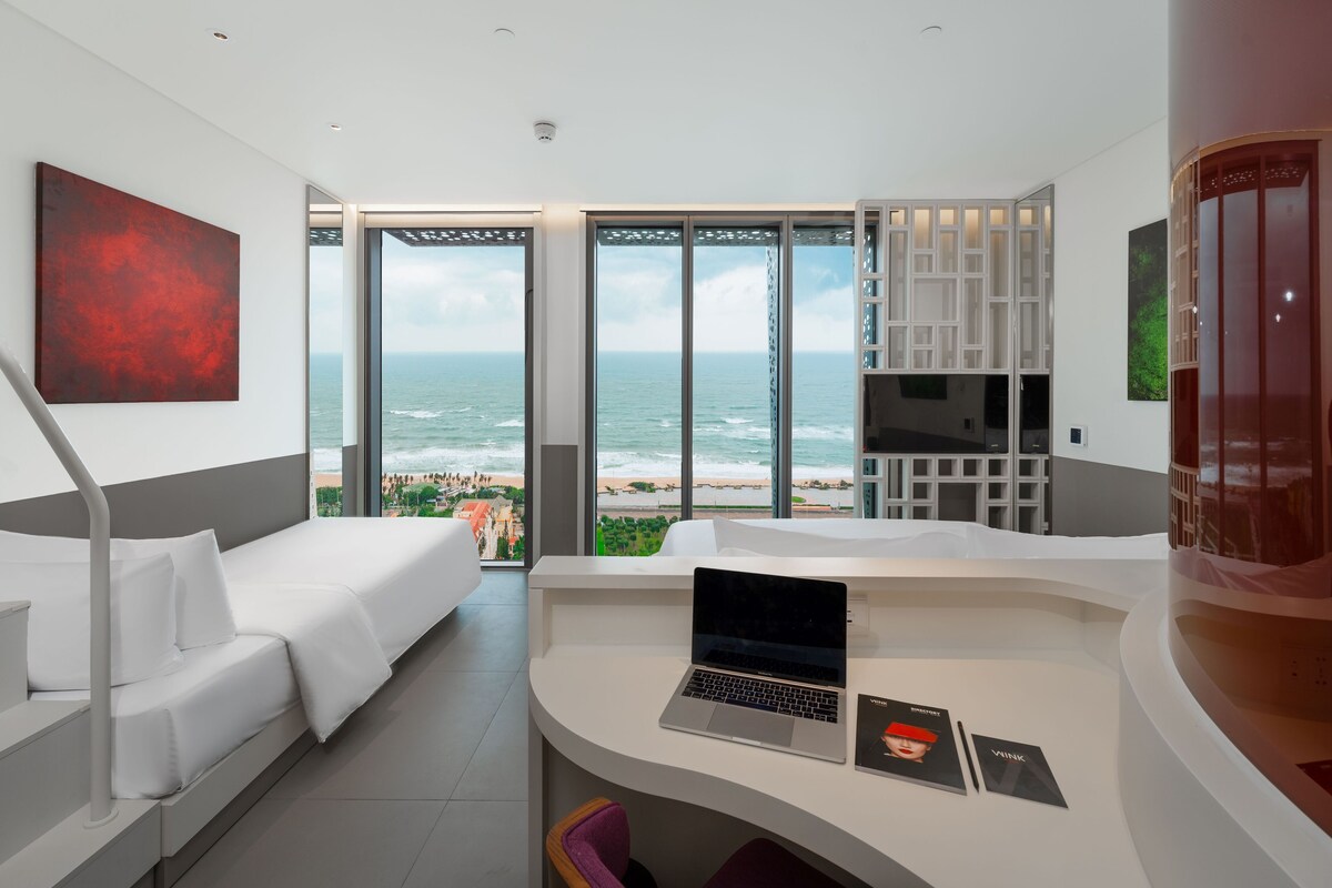 Une chambre d’hôtel moderne équipée d’un lit, d’un bureau avec un ordinateur portable et offrant une vue sur l’océan.