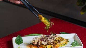 A hand using chopsticks to pick up a piece of food from a rectangular white plate on a red cloth.