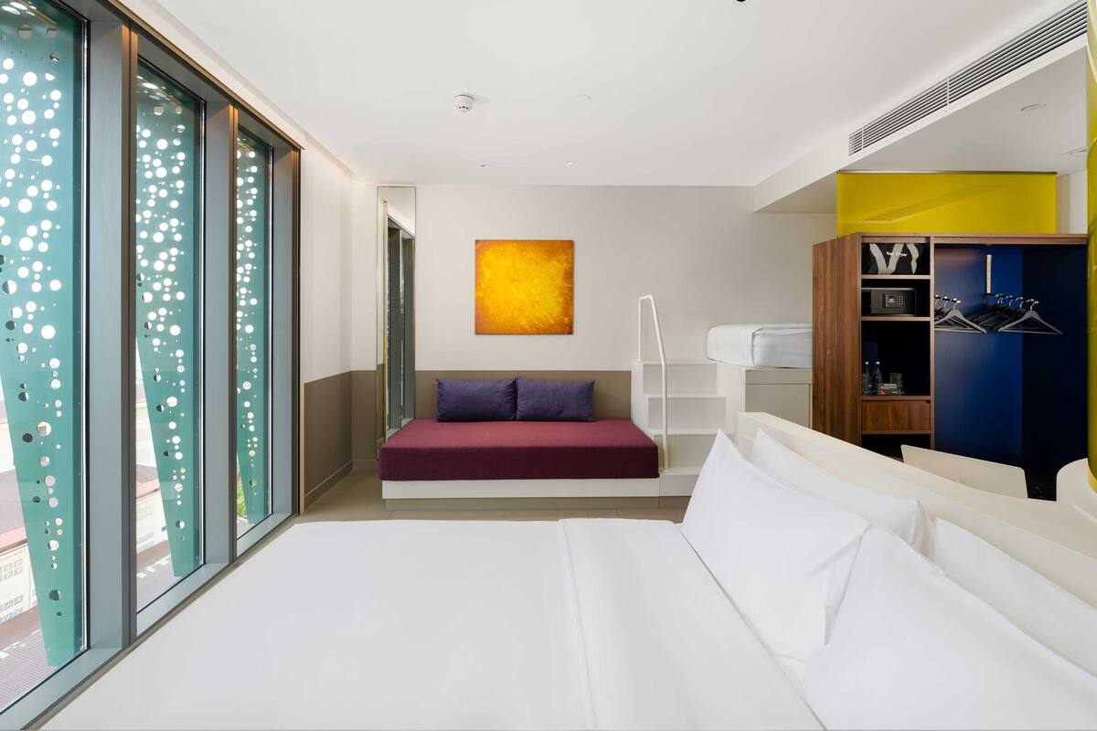 Une chambre d’hôtel moderne dotée d’un grand lit, d’un coin salon avec un coussin rouge, d’une armoire où sont suspendus des vêtements et d’une grande fenêtre offrant une vue dégagée.