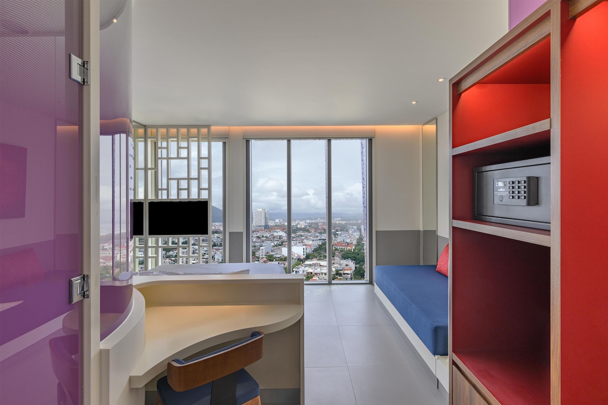 Ein modernes Hotelzimmer mit einem großen Fenster, einem Flachbildfernseher, einem roten Bücherregal und einem blauen Sofa.