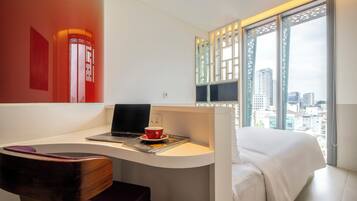 A modern hotel room with a desk, laptop, and a cup, a bed, and a large window with a city view.