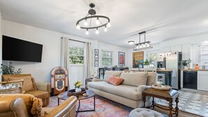 A living room with a sofa, armchair, coffee table, and ottoman. There is a television mounted on the wall, a radio, and a chandelier. The room has a large window with curtains. - The Juke Joint: Game Room, Pet-Friendly BHM Home! (Birmingham)