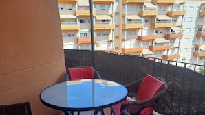 Mesa redonda con sillas de mimbre y cojines rojos en un balcón con vista a edificios de apartamentos.