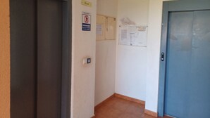 Un pasillo estrecho con una puerta cerrada a la derecha, un interruptor de luz y un tablón de anuncios con varios papeles y un cartel de prohibido fumar.