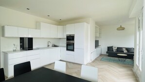 Una cocina moderna con armarios blancos, encimera negra y horno empotrado. Cuenta con un comedor con mesa y sillas negras, y un sofá de color oscuro en la sala.