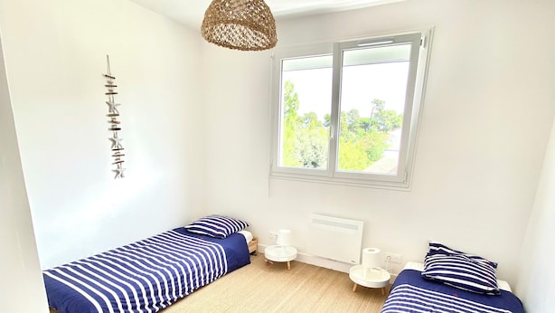 Una habitación pequeña y luminosa con dos camas, un ventanal grande y una lámpara colgante.