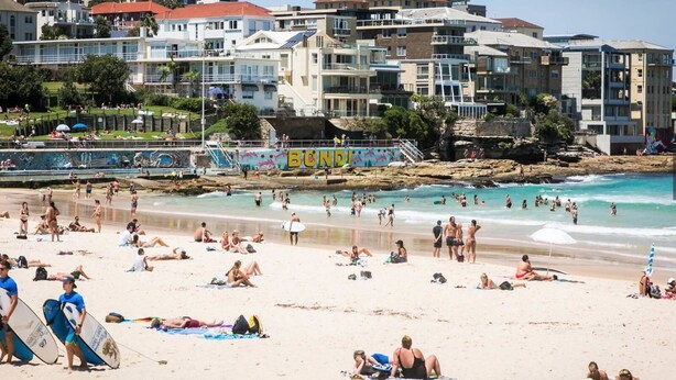 A crowded beach with people sunbathing and swimming, a lifeguard tower, and buildings in the background. - Stylish Sun Drenched North Bondi Beach Oasis (North Bondi)