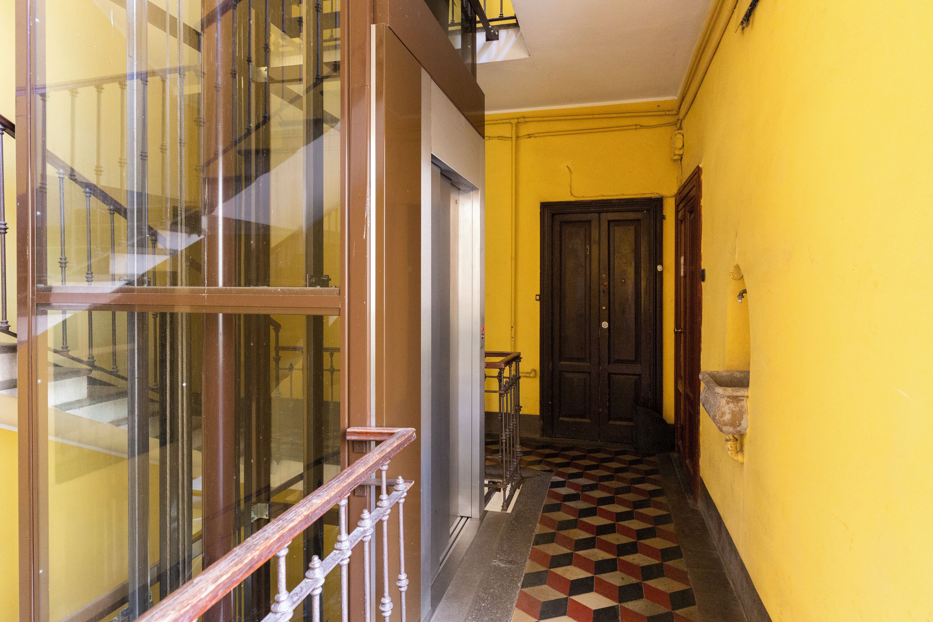 A hallway with a checkered floor, a wooden door, and a staircase with glass railings.