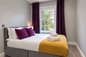 A neatly made bed with white and purple pillows, a mustard-colored throw blanket, and rolled towels on the side. - Stylish 3-Bedroom Apartment in Greater London (London)