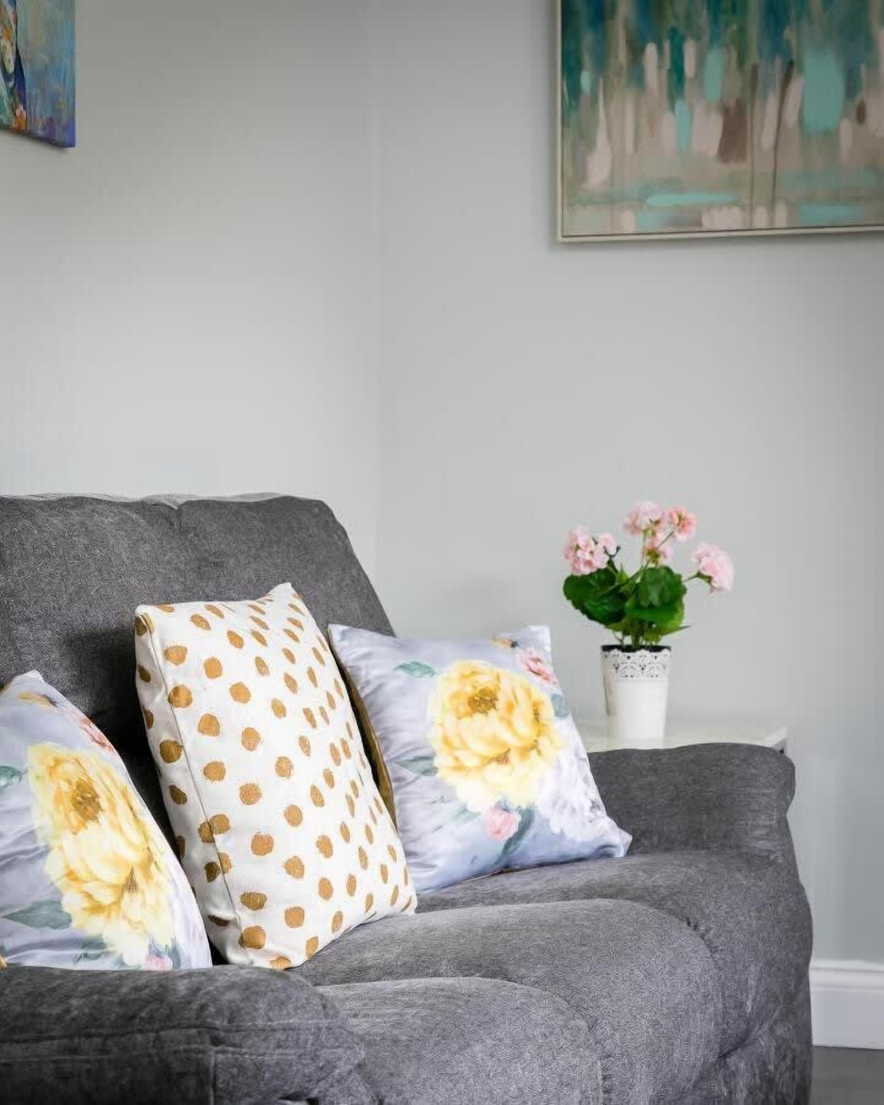 A grey sofa with two pillows, one with a polka dot pattern and the other with a floral design. A small white table with a potted plant is visible in the background.