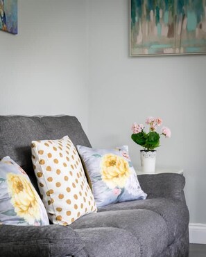 Ein graues Sofa mit zwei Kissen, eines mit Punkten und das andere mit Blumenmuster. Im Hintergrund ist ein kleiner weißer Tisch mit einer Topfpflanze zu sehen.