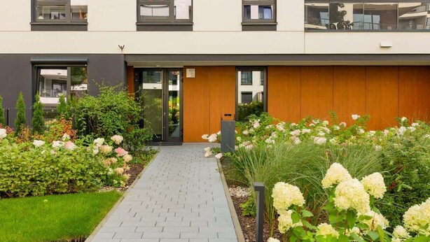 Un edificio moderno con entrada pavimentada, puerta de madera y jardín con diversas plantas y flores.
