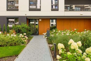 A modern building with a paved entrance, a wooden door, and a garden with various plants and flowers. - Stylish Apartment in Ochota | Parking, Balcony, Close to Airport (Warszawa)