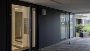 A modern building exterior with a glass door entrance, a tiled patio area, and a small planter with plants.