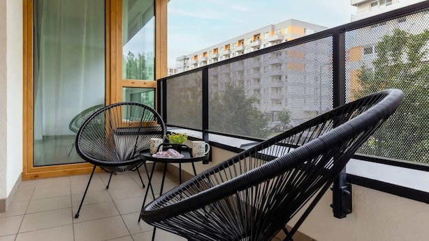 A balcony with two black wicker chairs, a small table with a bowl of fruit and a mug, and a large window with a view of a building.