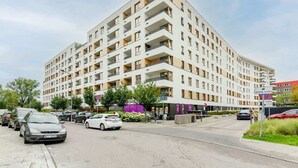 A multi-story hotel with a modern design, featuring balconies on each floor, a parking area in front, and a pedestrian walkway.