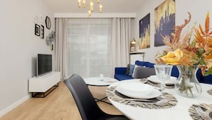 A modern living room with a dining area, featuring a flat-screen TV, a sofa, a chandelier, and a vase with flowers.