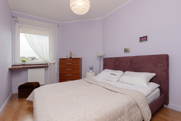 A bedroom with a bed, a dresser, a nightstand, a lamp, and a window with curtains.