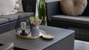 Un espace extérieur moderne, aménagé avec des sièges, une table en rotin foncé, une lampe en métal et un vase rempli de fleurs.