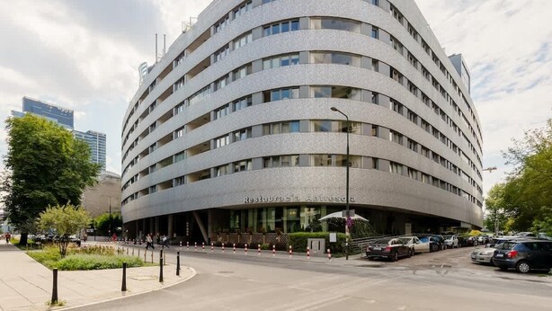 Un immeuble à plusieurs étages doté d’une façade incurvée, d’une entrée abritée et d’un parking où sont garées des voitures.