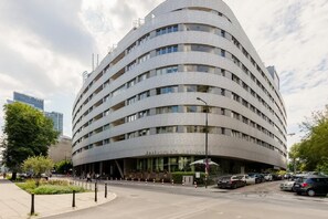 Un immeuble à plusieurs étages doté d’une façade incurvée, d’une entrée abritée et d’un parking où sont garées des voitures.