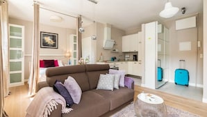 Moderni hotellihuone, jossa on sohva, pieni pyöreä pöytä, keittiöalue ja kylpyhuone, jossa on matkalaukku.