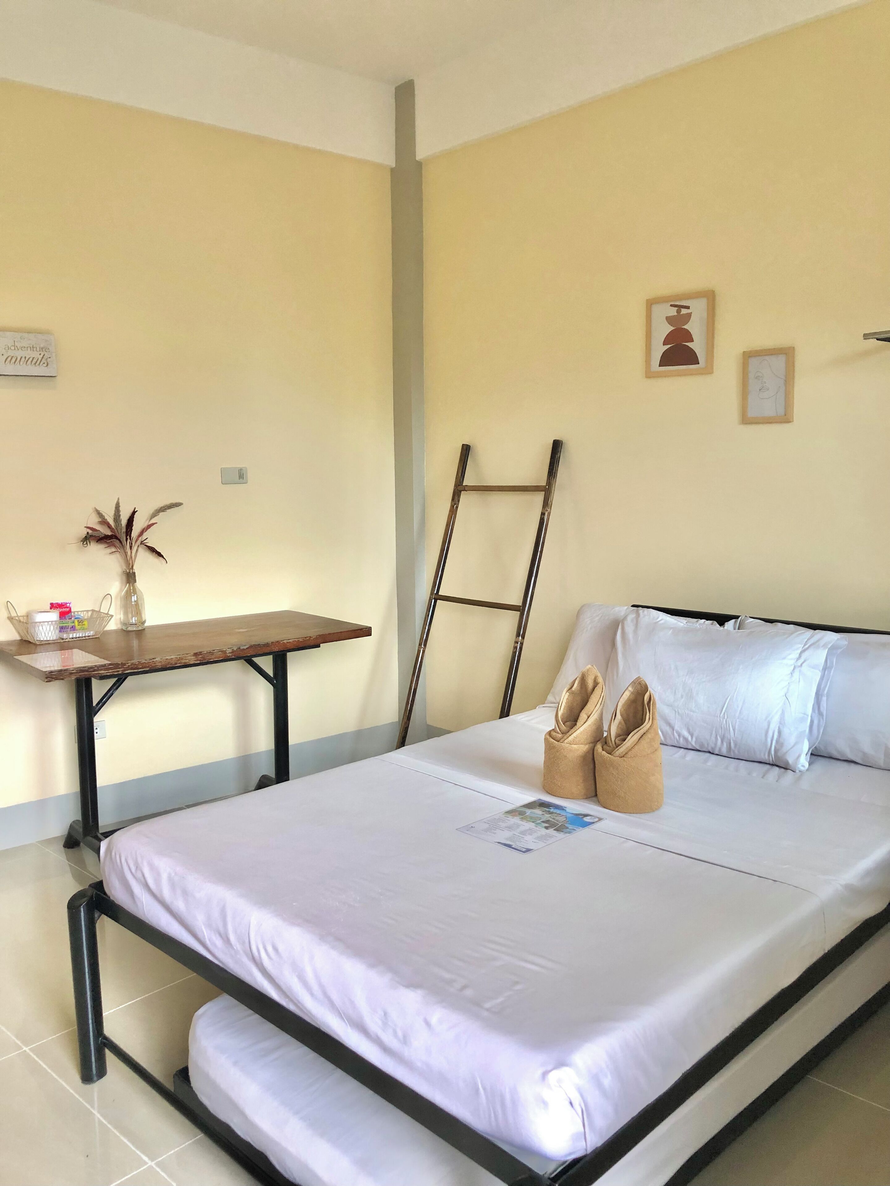 Un letto singolo con materasso e cuscini bianchi, un comodino in legno con un vaso e un cesto, e una scala appoggiata al muro.