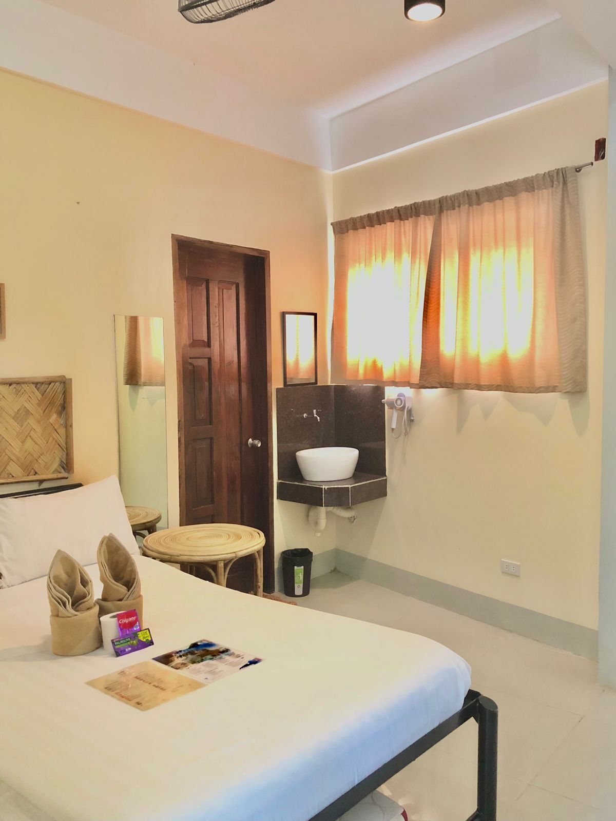 A hotel room with a bed, a small table, a chair, a wooden door, a window with curtains, a wall-mounted sink, and a trash bin.