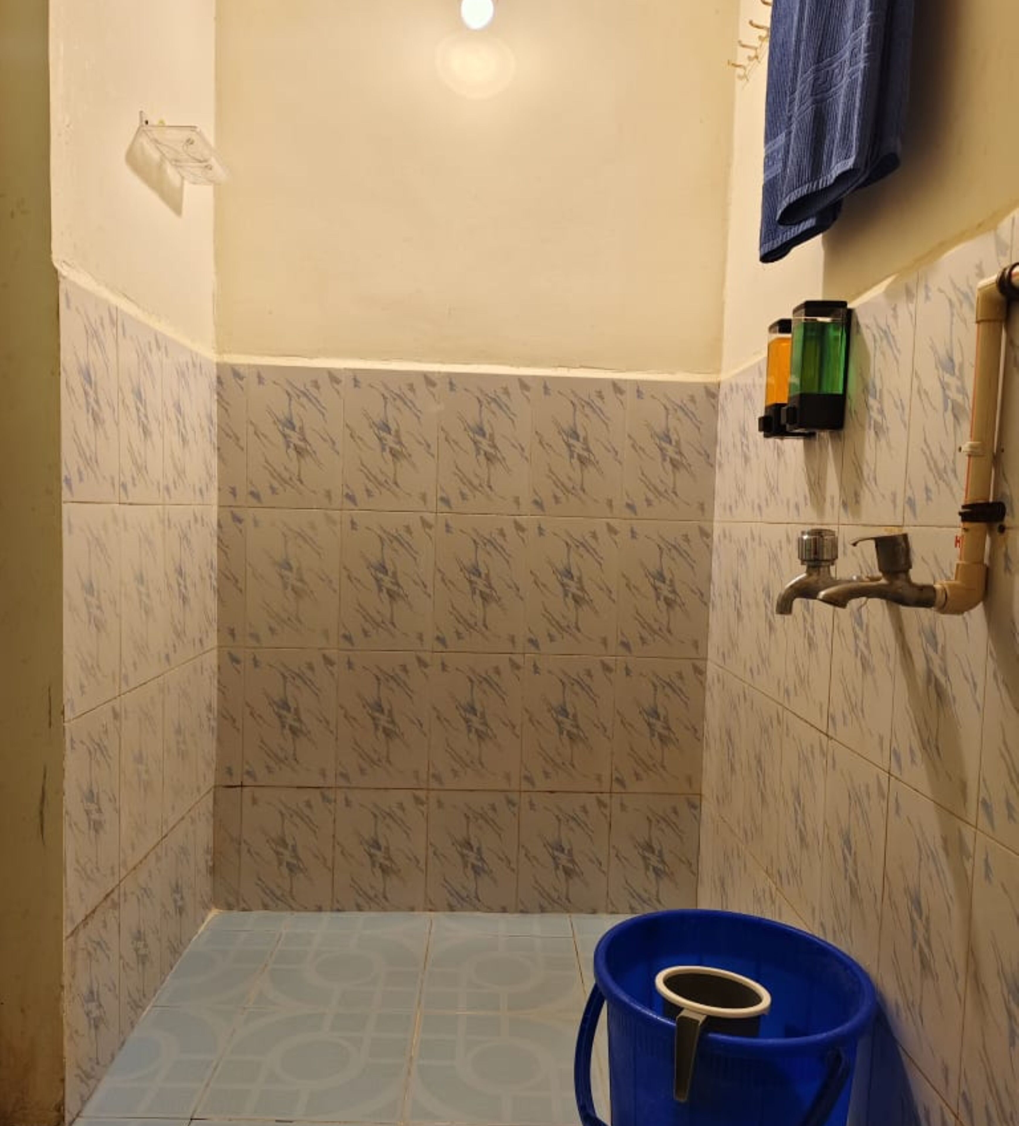 A bathroom with a shower area, a towel rack, a soap dispenser, and a blue bucket.
