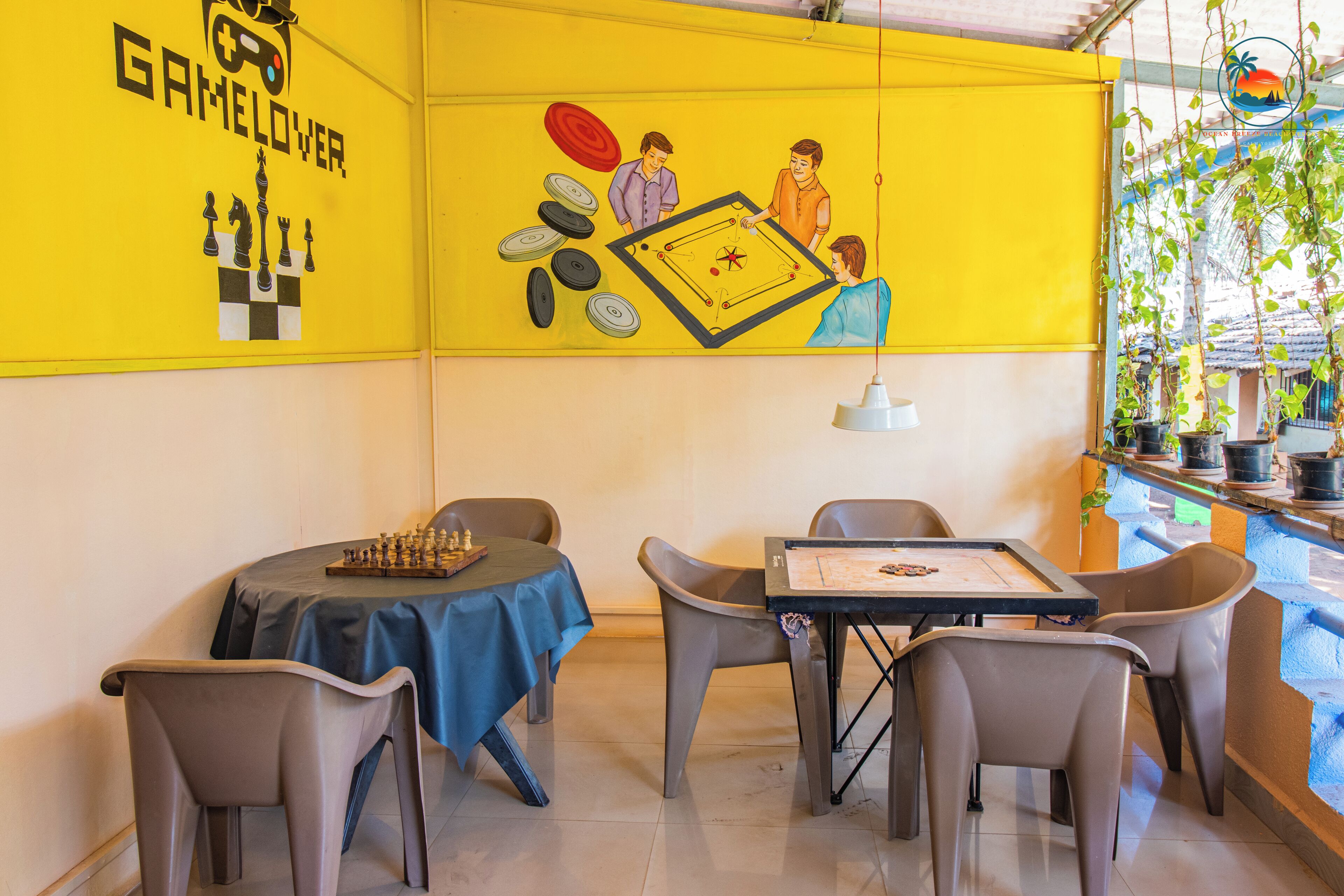 A room with a yellow wall featuring a mural of people playing pool, a blue table with a chessboard, and a small table with a board game.