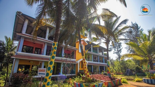 Un edificio de dos pisos con un letrero de dormitorio, rodeado de palmeras y coloridos macizos de flores.