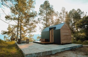 A small cabin with a sloped roof, a wooden exterior, and a concrete platform with a bench. - Bobocabin Signature Toba (Ajibata)