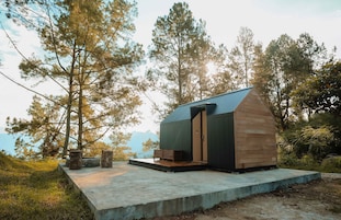 Uma cabana pequena com telhado inclinado, fachada de madeira e um alpendre de concreto com um banco.
